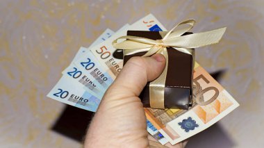 Bir adam içinde dolar banknotları olan bir hediye kutusu tutuyor, yakın plan. Sevgiliye hediye, tatil konsepti, kadınlar günü, sevgililer günü, doğum günü. Elinde kurdeleler ve 100 dolarlık banknotlar olan kahverengi kutu.