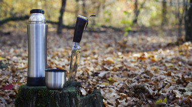 Termos ve av bıçağı. Turizm, hayatta kalma ve sonbaharda ormanda avlanma kavramı. Bir termos çay, bir fincan ve bir bıçak, eski bir ağaç kütüğünün üzerinde. Parktaki kuru yapraklar. doğada dinlen..