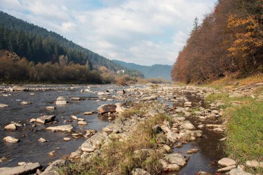 dağlar, taştan sahil, dağ nehri, sonbahar ormanı ve bulutlu gökyüzü. Kayalık kıyıları olan dağ nehri. Dağlarda güneşli bir gün. Batı doğası. Turizm, Karpatlara seyahat.