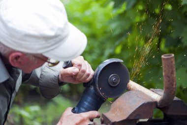 Yaşlı adam çalışıyor. Metal bir boruyu kesmek. Kıvılcım. Boru mengeneye sıkışmış. Adamın elleri öğütücü tutuyor. Eski demir mengene, paslı boru. Metalle çalışan elektrikli bir alet. Bulgarca, disk öğütücü