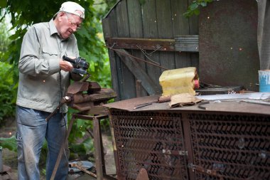 Yaşlı adam çalışıyor. Metal bir boruyu kesmek. Boru mengeneye sıkışmış. Adamın elleri Bulgar bir disk öğütücüsünü tutuyor. Eski demir mengene, paslı boru. Metalle çalışan elektrikli araç.