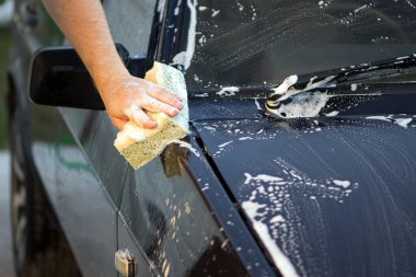 Bir el bezi ve sabun köpüğüyle arabanın kaputunu yıkar. Adam araba kaputunu yıkama bezi ve şampuanla yıkıyor, araba köpüğüyle kaplı, araba yıkama servisi var. Siyah yüzeyi temizlemek