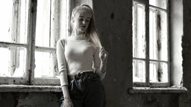 Genç kız eski pencerenin yanında duruyor. 18 yaşında, şık bir genç, eski bir ev, kırık camlar, güzel genç bir kız. Duygular, yaşam tarzı. Yakın plan. Saç düzleştirir, saç stili yapar. yıkılan bina, savaş zamanı