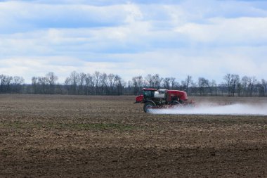 Bulutlu bir günde tarlayı sulayan sulama sistemi olan kırmızı traktör. Tarımsal makineler, tarlada çalışmak. Mevsimlik iş. Traktör toprağı gübreler.