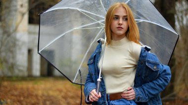 Sonbahar parkında şemsiyeli genç bir kız yürüyor. Modern kız şehir parkında yürüyor. Mevsimlik stil, sonbahar güzelliği. Sonbahar mevsimi, mavi ceketli, şeffaf şemsiyeli, sonbahar yapraklı.