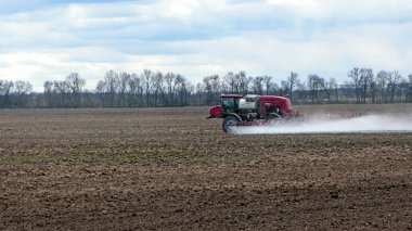 Bulutlu bir günde tarlayı sulayan sulama sistemi olan kırmızı traktör. Tarımsal makineler, tarlada çalışmak. Mevsimlik iş. Traktör toprağı gübreler. Sahada küçük traktörler.