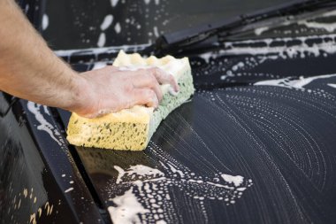 Bir el bezi ve sabun köpüğüyle arabanın kaputunu yıkar. Adam araba kaputunu yıkama bezi ve şampuanla yıkıyor, araba köpüğüyle kaplı, araba yıkama servisi var. Siyah yüzeyi temizlemek