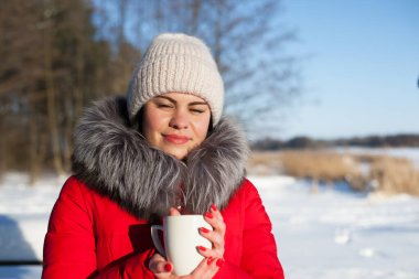 Genç bir kadınla kış ormanında çekilmiş bir fotoğraf. Karlı bir parktaki kız. Elinde bir fincanla kırmızı bir ceketle. Doğada kış yürüyüşü. Soğuk mevsim, güneşli gün. Portre, çay içmek. yakın plan