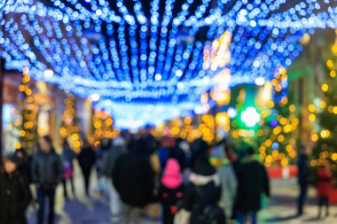 Bir grup insan Noel ışıklarıyla caddede yürüyorlar. Işıklar parlak ve renkli, neşeli bir atmosfer yaratıyor. İnsanlar kalın giyinmiş, bazıları şapka takıyor.