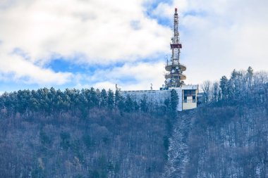 Uzakta kırmızı tepeli uzun bir kule görülüyor. Gökyüzü bulutlu ve ağaçlar çıplak.