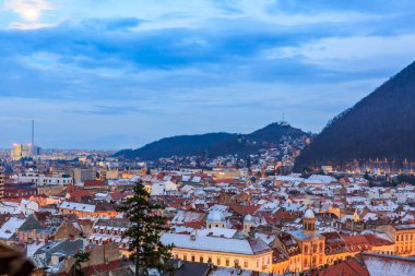 Önünde büyük bir ağaç ve arkasında bir dağ olan bir şehir manzarası. Gökyüzü bulutlu ve güneş batıyor.