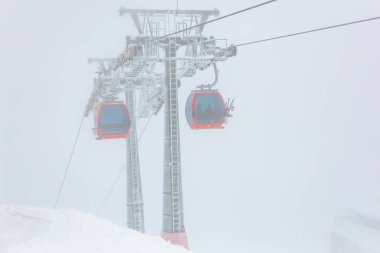 Sisli bir kış gününde, karlı dağ manzarasında kırmızı teleferikler..