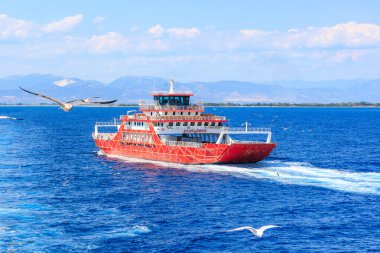 Kırmızı bir tekne arka planda uçan bir kuşla okyanusta gidiyor. Gemi yolcuları taşıyor ve kıyıya doğru gidiyor. 5 Ağustos 2024 Keramoti 'den Yunanistan' a feribot