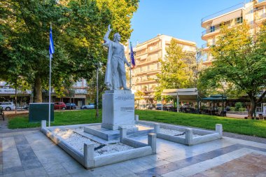 Parktaki bir çeşmenin önünde duran bir adam heykeli. Heykel beyaz bir taş leğen ve birkaç bayrakla çevrili. 7 Ağustos 2024 Yunanistan