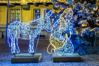 İsa gibi giyinmiş bir adam bankta bir eşeğin yanında oturur. Eşek de ışıklandırılmış ve o da Noel süslemesinin bir parçası. 13 Aralık 2024 Brasov Romanya