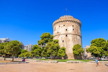 Üzerinde bayrak olan uzun bir kule bir şehrin önünde duruyor. Gökyüzü açık ve mavidir ve bölgede yürüyen birkaç kişi vardır. 8 Ağustos 2024 Selanik Yunanistan