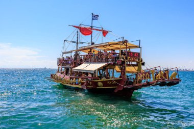 Kırmızı yelkenli ve beyaz tenteli büyük bir tekne okyanusta yüzüyor. Tekne insan kaynıyor ve teknede birkaç bayrak var. 8 Ağustos 2024 Selanik Yunanistan