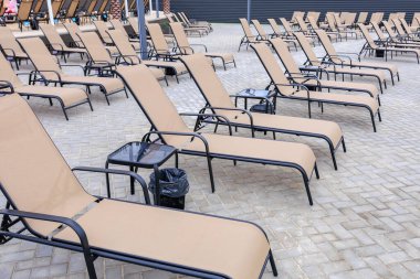 Tuğla bir verandada bir sıra bronzlaşmış şezlong dizilmiştir. Sandalyeler boş ve yakınlarda bir çöp kutusu var. Sahne rahatlatıcı ve sakin bir hava veriyor.