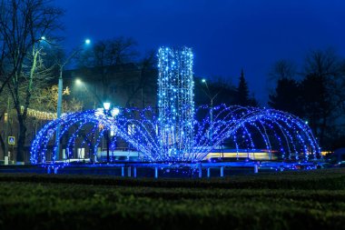 Geceleri ağaçların ve sokak lambalarının arasında açık mavi bir çeşme. 20 Aralık 2024 Balti Moldova.