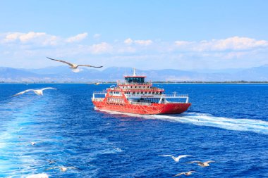 A red boat is sailing on the ocean with a flock of seagulls flying in the background. August 5, 2024 Ferry from Keramoti to Thassos island Greece clipart