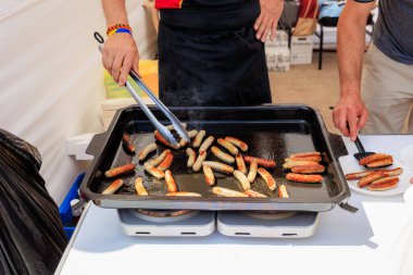 Kapalı çekimde geniş bir tavada sosisleri ızgarada pişirirken sosyal bir olayı gündelik bir yemek sahnesiyle vurguluyor..