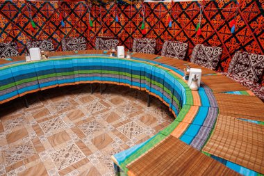 Renkli masa örtüsü ve çeşitli sandalyeleri olan büyük yuvarlak bir masa. Masa renkli bir duvarı ve halısı olan bir odayla çevrili. Kırgız otantik göçebe yurdunda kutlama masası.