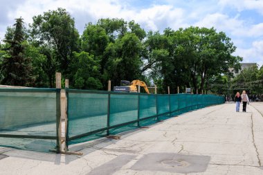 İnşaat alanı yeşil bariyerli çitlerle çevrili ve şehir merkezinde bulutlu gökyüzü olan ağaçlar var. 29 Haziran 2023 Beltsy Moldova.