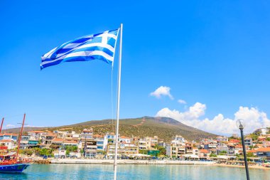 Bir kasabanın üzerinde mavi ve beyaz bir bayrak dalgalanıyor. Bayrak bir direğin üzerinde ve sudan görülebiliyor. 6 Ağustos 2024 Thassos Adası Yunanistan