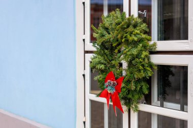 Pencerede kırmızı kurdeleli ve çam iğneli güzel bir çelenk asılı. Çelenk kırmızı kurdele ve çam iğneleriyle süslenmiş ve mavi arka planı olan bir pencereye asılmış.