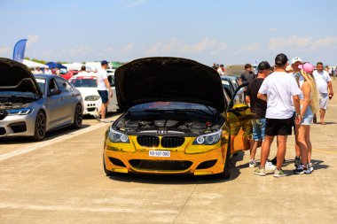 A shiny gold chrome BMW with the hood open, surrounded by people observing and checking details on the vehicle. July 2, 2023. Liadoveni, Moldova. clipart