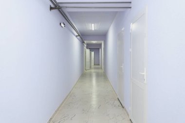 Açık kapıları ve soluk mavi duvarları olan uzun, boş bir koridor..
