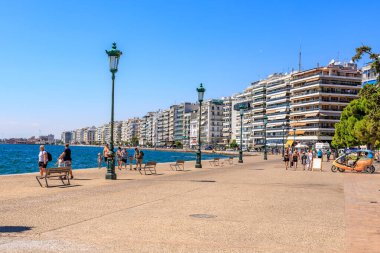 İnsanların banklarda yürüyüp oturduğu bir plaj sahnesi. Sahil yüksek binaları olan bir şehre yakındır. 8 Ağustos 2024 Selanik Yunanistan