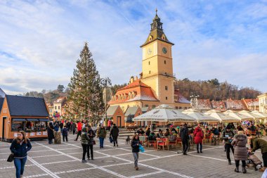 Büyük bir Noel ağacı ve saat kulesi olan işlek bir şehir meydanı. İnsanlar etrafta dolanıp masalarda oturuyorlar. 14 Aralık 2024 Brasov Romanya