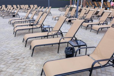 Tuğla bir verandada bir sıra bronzlaşmış şezlong dizilmiştir. Sandalyeler boş ve yakınlarda bir çöp kutusu var. Sahne rahatlatıcı ve sakin bir hava veriyor.