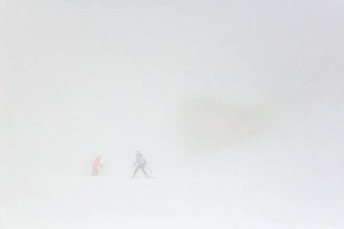 İki kayakçı yoğun kar yağışı altında sisli dağ yamacında yol alırlar..