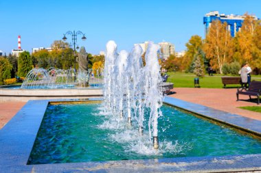 Parkta dört su borusu olan bir çeşme var. Su havaya fışkırıyor, serin ve ferahlatıcı bir atmosfer yaratıyor. Park, insanların oturabileceği bir yer sağlayan banklarla dolu.