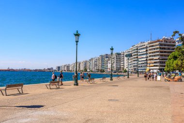 İnsanların banklarda yürüyüp oturduğu bir plaj sahnesi. Sahil yüksek binaları olan bir şehre yakındır. 8 Ağustos 2024 Selanik Yunanistan