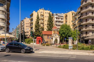 Beyaz çatılı küçük kırmızı bir bina büyük bir binanın önünde duruyor. Arkasındaki büyük bina yüksek bir apartman, kilise. 8 Ağustos 2024 Selanik Yunanistan