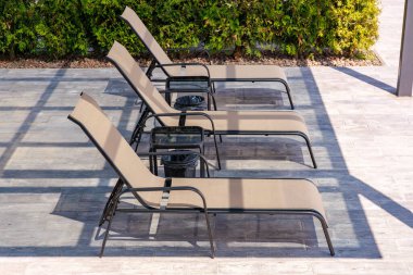 Üç şezlong terasta oturuyor. Sandalyeler bronzlaşmış ve sıraya dizilmişler. Sandalyelerin ortasında bir bank var.