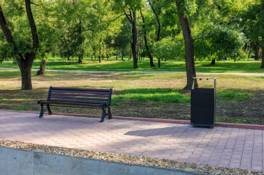 Bir park bankı kaldırımda bir çöp kutusunun yanında oturur. Tezgah boş ve çöp kutusu dolu. Park sessiz ve huzurlu, ağaçlar gölge ve sükunet sağlıyor.