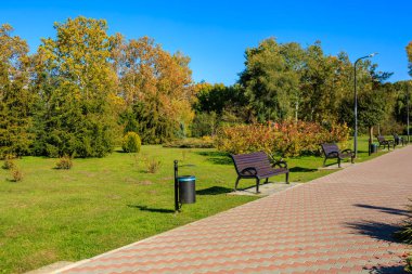 Birkaç bank ve çöp tenekesi olan bir park. Park boş ve gökyüzü mavi.