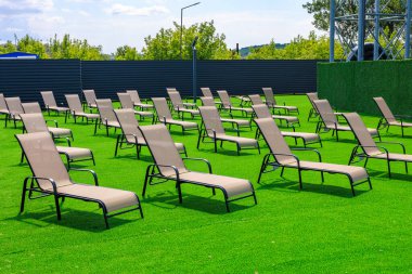 Siyah çitleri ve etrafa saçılmış sayısız şezlongları olan geniş çimenli bir alan. Sandalyeler boş, huzurlu ve rahatlatıcı bir atmosfer izlenimi veriyor.