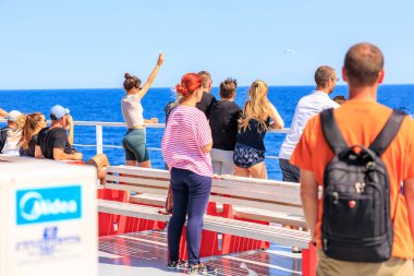 Bir grup insan bir teknede durup okyanusu izliyor. Pembe gömlekli bir kadın bankta duruyor, suya bakıyor. 5 Ağustos 2024 Keramoti 'den Yunanistan' a feribot