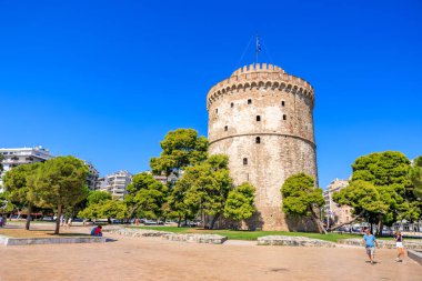 Üzerinde bayrak olan uzun bir kule bir şehrin önünde duruyor. Gökyüzü açık ve mavidir ve bölgede yürüyen birkaç kişi vardır. 8 Ağustos 2024 Selanik Yunanistan