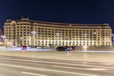 Şehir meydanında gece vakti aydınlatılmış camları ve arabaları olan tarihi bir bina. 8 Mart 2025 Bükreş Romanya.