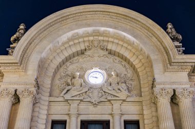 Gece gökyüzünün altındaki saat ve heykellerle dekore edilmiş mimari cephe.