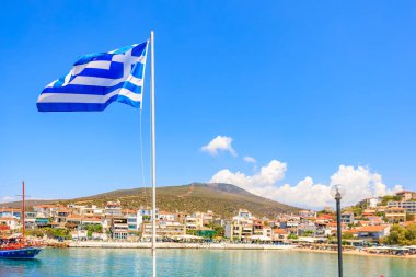 Bir kasabanın üzerinde mavi ve beyaz bir bayrak dalgalanıyor. Bayrak direği suyun içinde. 6 Ağustos 2024 Thassos Adası Yunanistan