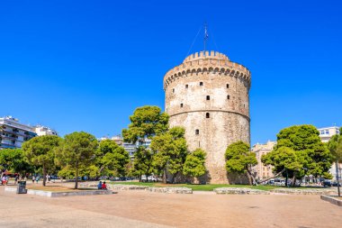 Üzerinde bayrak olan uzun bir kule bir şehrin önünde duruyor. Gökyüzü açık ve mavidir ve bölgede yürüyen birkaç kişi vardır. 8 Ağustos 2024 Selanik Yunanistan