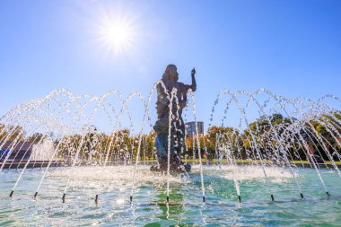 Bir insan heykeli çeşmenin önünde duruyor. Çeşme havaya su püskürtüyor. Güneş ışıl ışıl parlıyor, sahneyi parlak ve neşeli gösteriyor. 27 Ekim 2024 Chisinau Moldova