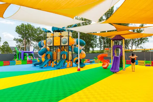 Büyük sarı ve beyaz şemsiyeli bir oyun alanı. Bir kadın çocuk parkında yürüyor. Çocuk parkında oynayan bir sürü çocuk var. 8 Haziran 2024 Balti Moldova